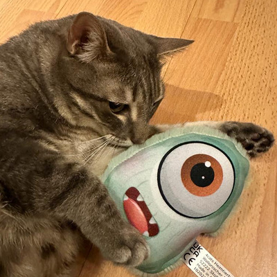 Liegende Katze hält Katzenkissen im Design grünes Monster in beiden Pfoten und schnuppert daran